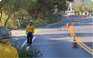 Road crew and fire-safety workers stand along a two-lane road lined with traffic cones, clearing vegetation and assessing an emergency egress route between Rancho Road and Galbreth Road.