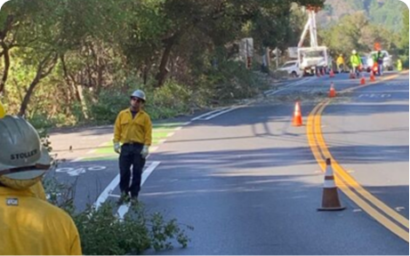 Road crew and fire-safety workers stand along a two-lane road lined with traffic cones, clearing vegetation and assessing an emergency egress route between Rancho Road and Galbreth Road.